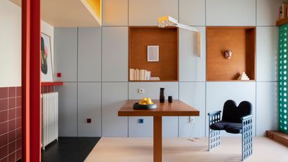 A dining area with wall-to-wall built-in cabinetry in pale blue, with a fold-down wooden table, red steel column and terracotta nooks